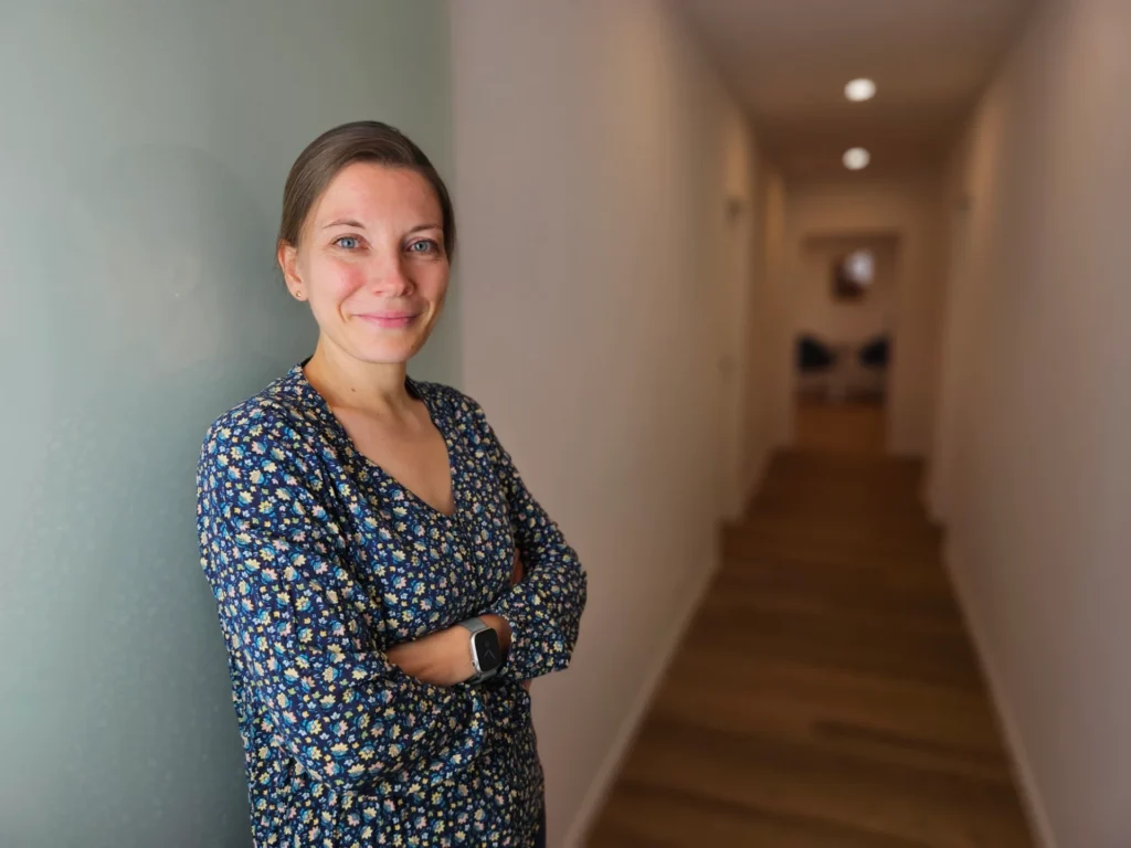 Portrait einer Frau mit dunkelblauem Blumenmuster-Kleid, lächelnd, die Arme verschränkt an einer grünlich gestrichenen Wand in einem hellen Flur stehend.