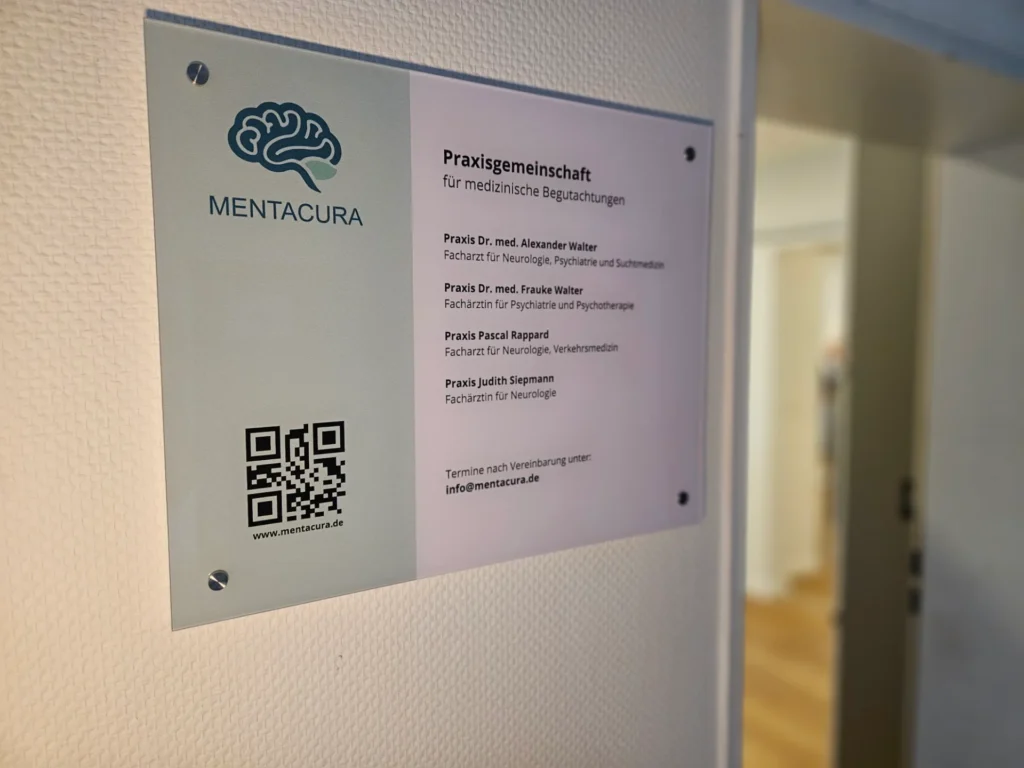 Mentacura-Plakat an einer hellen Wand mit Gehirn-Logo, Text zur Praxisgemeinschaft für medizinische Begutachtungen, QR-Code und Website www.mentacura.de