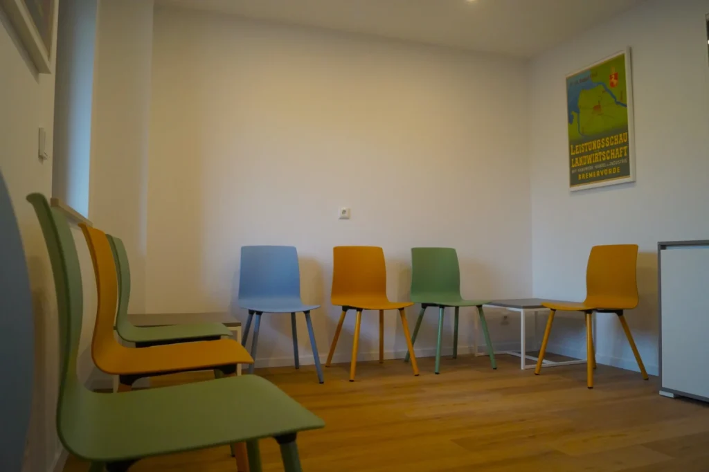 Warteraum mit bunten Stühlen in einem schlichten Raum, Holzfußboden, weiße Wände und ein farbiges Poster an der rechten Wand, ruhige und einladende Atmosphäre