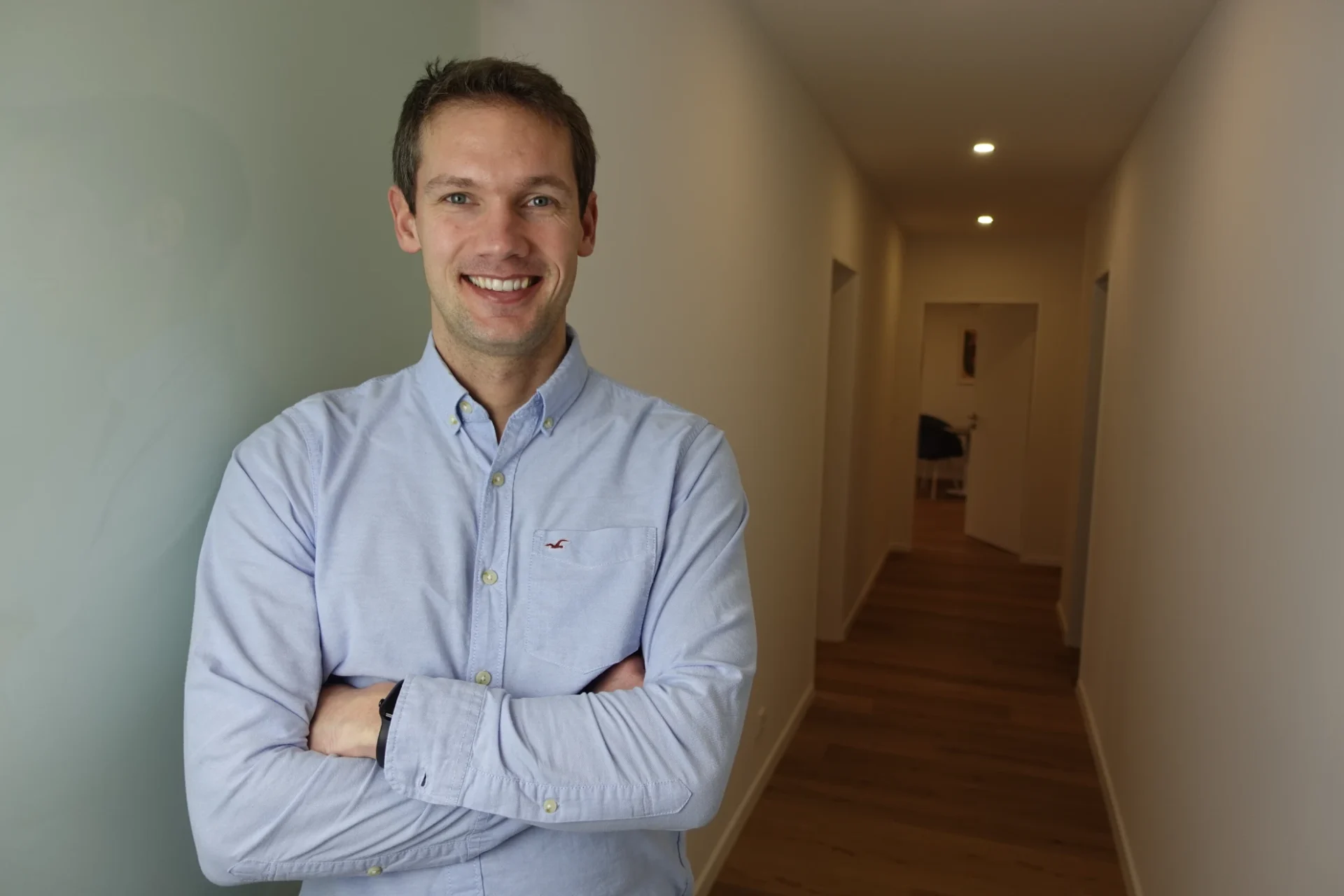 Mann in hellblauem Hemd lehnt sich an eine helle Wand in einem langen Flur, lächelt freundlich in die Kamera, Umfeld wirkt praktisch hell und einladend.